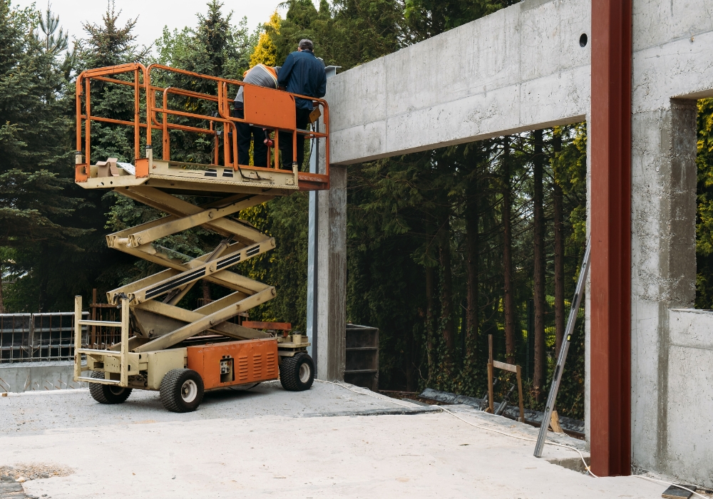 Deux personnes sur une petite nacelle orange. Elles se trouvent sur un chantier, probablement celui d'un bâtiment.