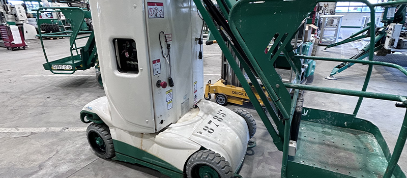 Une nacelle élévatrice de petite taille, blanche et verte, stationnée dans un entrepôt industriel. Cet équipement compact est conçu pour faciliter les travaux en hauteur dans des espaces restreints.