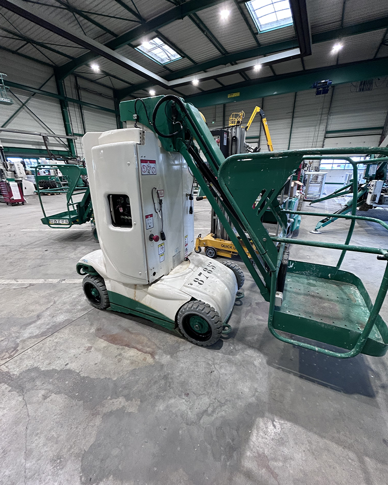 Une nacelle élévatrice de petite taille, blanche et verte, stationnée dans un entrepôt industriel. Cet équipement compact est conçu pour faciliter les travaux en hauteur dans des espaces restreints.