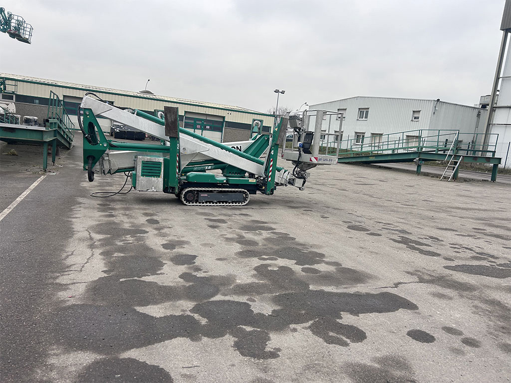 Une nacelle à l'arrêt et pliée, de couleur verte et blanche. Il semblerait qu'elle soit en attente de location sur le parking d'un entrepôt.