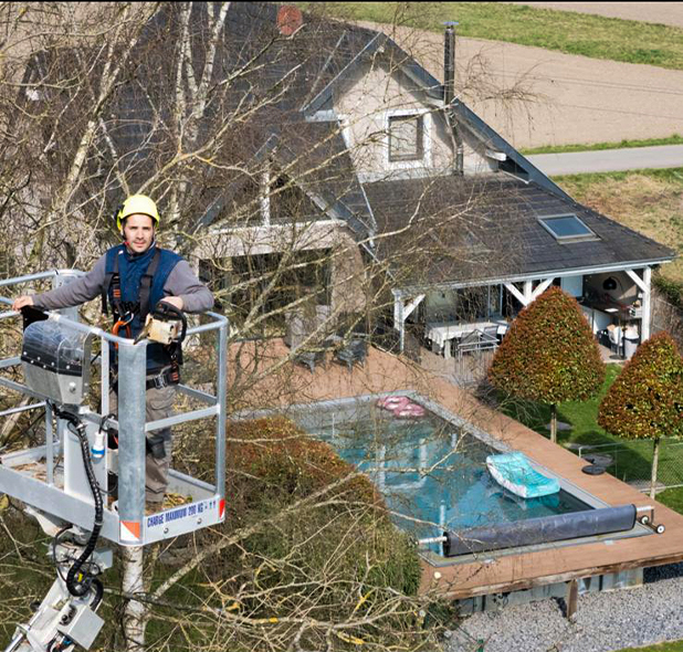 Un opérateur équipé d’un harnais et d’une tronçonneuse effectue un travail d’élagage en hauteur à l’aide d’une nacelle élévatrice devant une maison avec piscine.